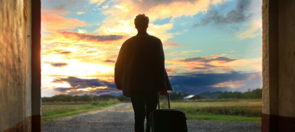 A traveller silhouetted in an archway at sunset, pulling a single wheeled suitcase.