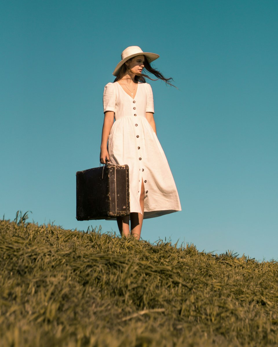 A woman in a pale dress standing on a hilltop against a deep blue sky, holding a vintage suitcase.