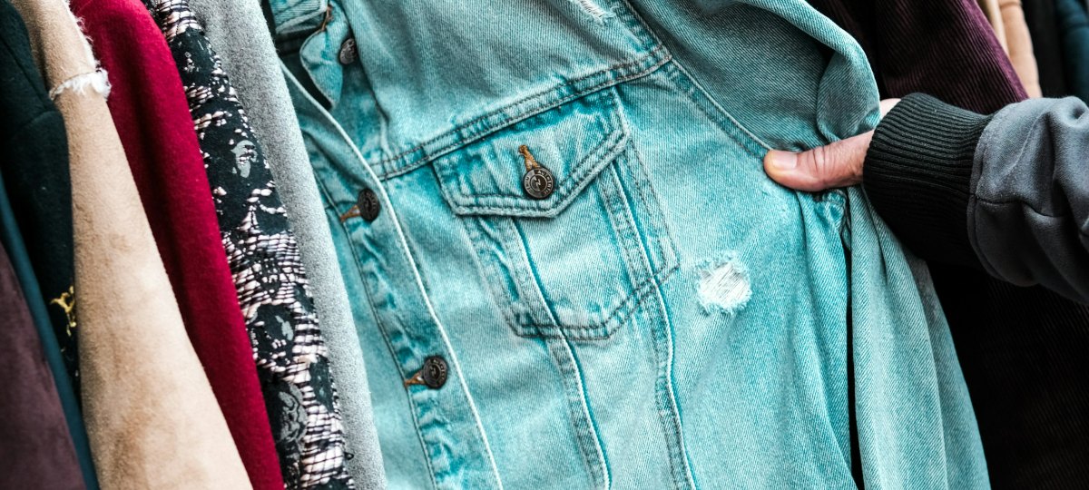 A hand reaching into a packed rail of second-hand jackets, holding open a faded pale-blue denim jacket among suede, wool and corduroy coats.