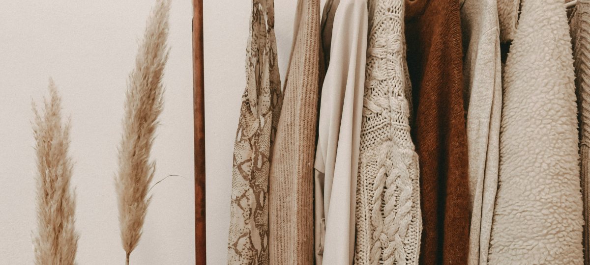 A rail of neutral knitwear and coats on white hangers beside dried pampas grass.