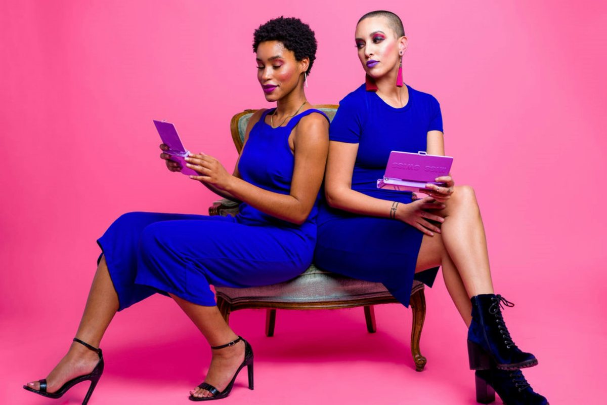 Two women in cobalt blue dresses seated back to back against a hot pink background.