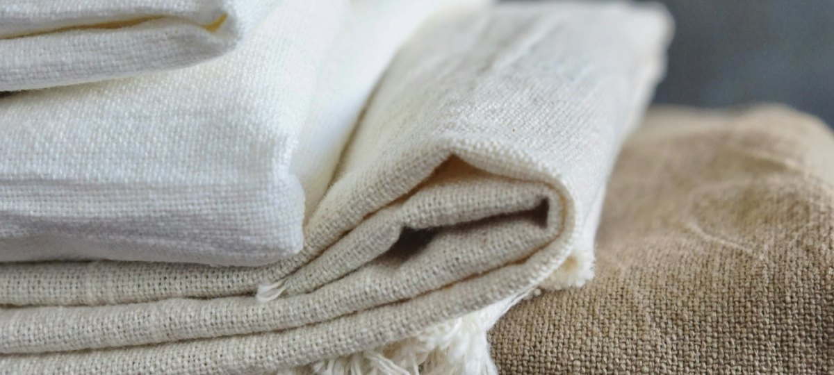 A folded stack of undyed natural textiles — loose-weave cream cotton with a fringed edge, on top of coarser natural linen — resting on a wooden bench.