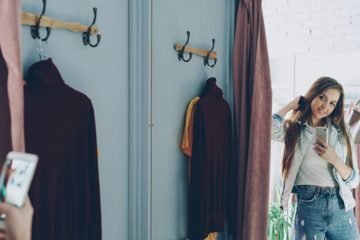 A woman photographing herself in a fitting-room mirror beside a rail of clothes.