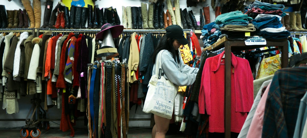 A woman in a cap and grey sweatshirt browsing a densely packed vintage shop, with rails of coats and knitwear, a row of leather boots on a high shelf and a stand of belts beside her.