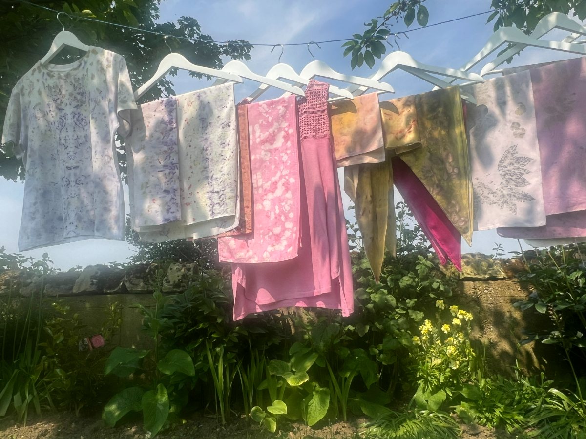Eco-printed cotton tops and a pink dress on white hangers against a blue sky, with garden plants below.