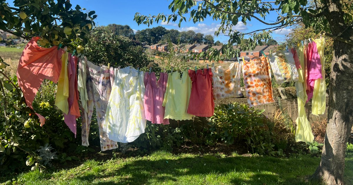 Plant-dyed clothes in coral, yellow, pink and lilac hanging on a washing line between an apple tree and a cherry tree, with rooftops and fields behind.