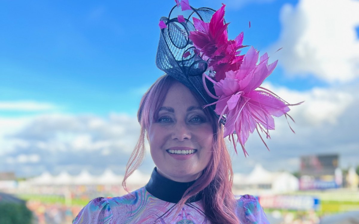Estelle Pearce at Doncaster Racecourse in a navy and pink feathered fascinator of her own design, smiling against a bright blue sky with the grandstand tents behind her.