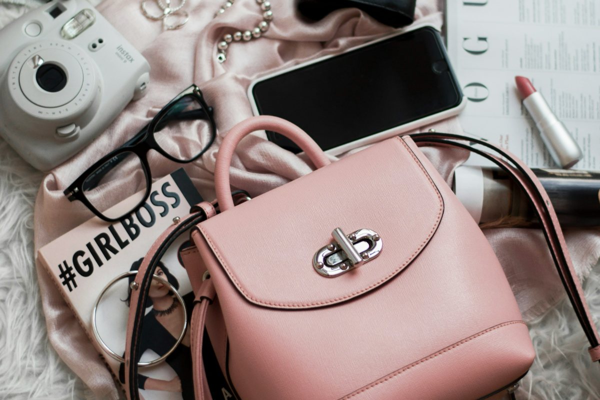 A flat lay of a pink handbag, glasses, lipstick, camera and magazines on a fur throw.