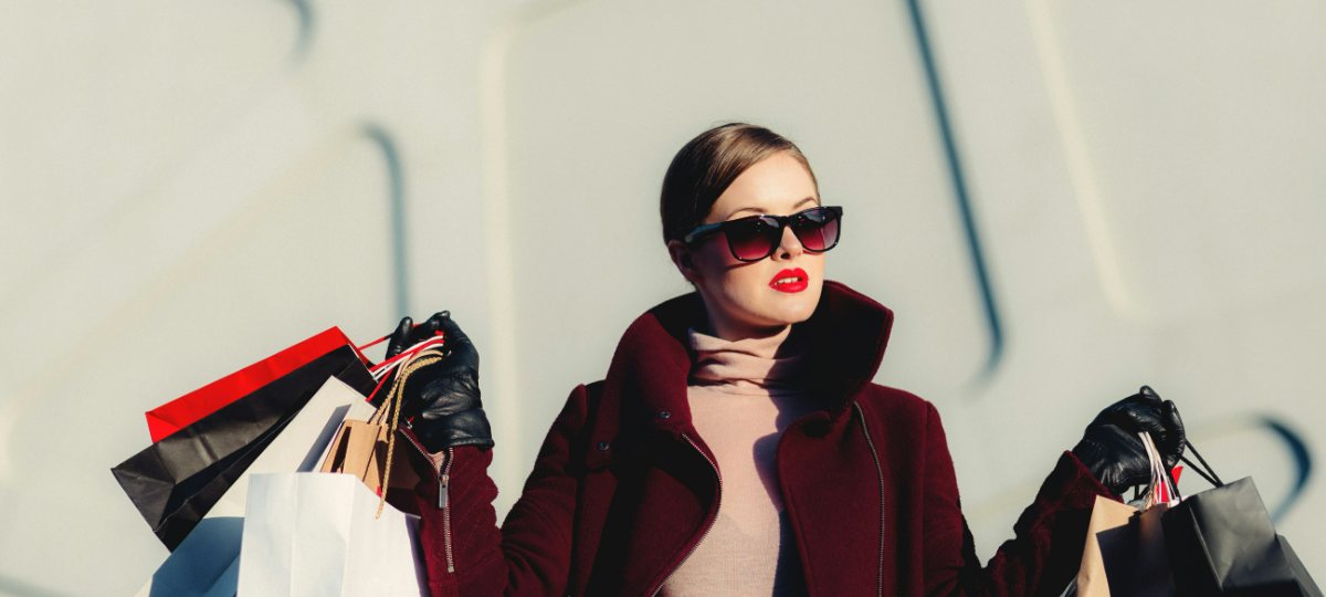 A woman in a burgundy coat and dark sunglasses carrying several paper shopping bags in both hands.