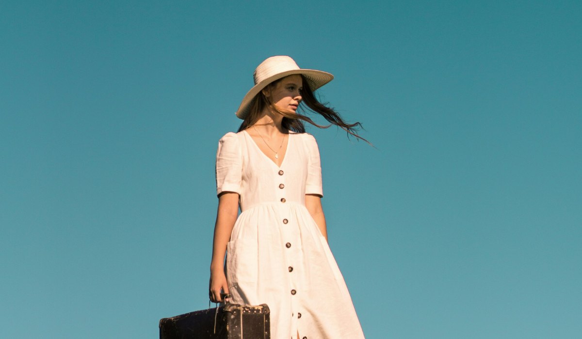 A woman in a cream linen button-through dress and straw sun hat standing on a grassy hill against a deep blue sky, holding an old leather suitcase.