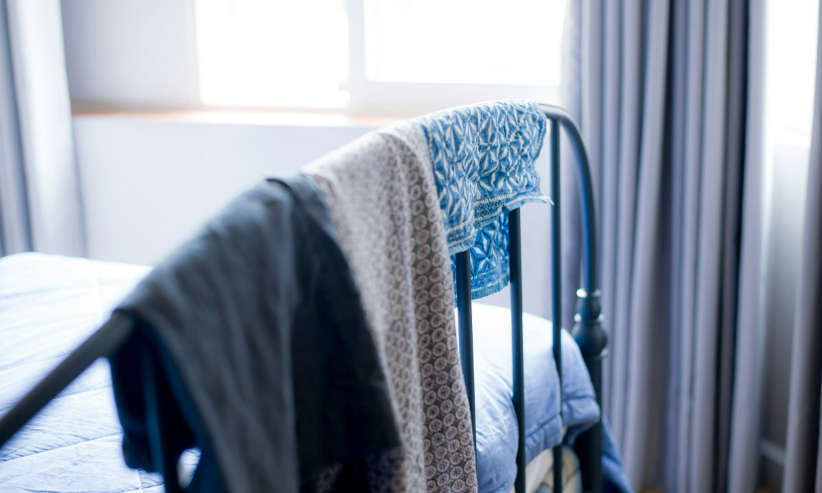 Three garments — a dark jacket, a patterned scarf and a blue print — draped over an iron bed frame by a bright window.