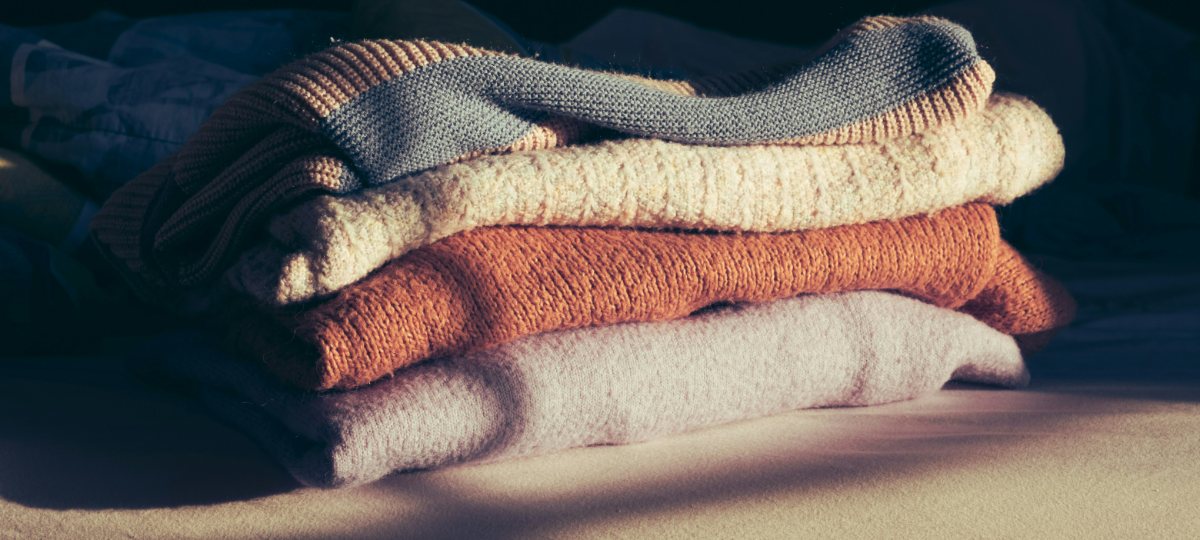 A stack of folded jumpers in grey, cream, rust and lilac, lit by low autumn sunlight.