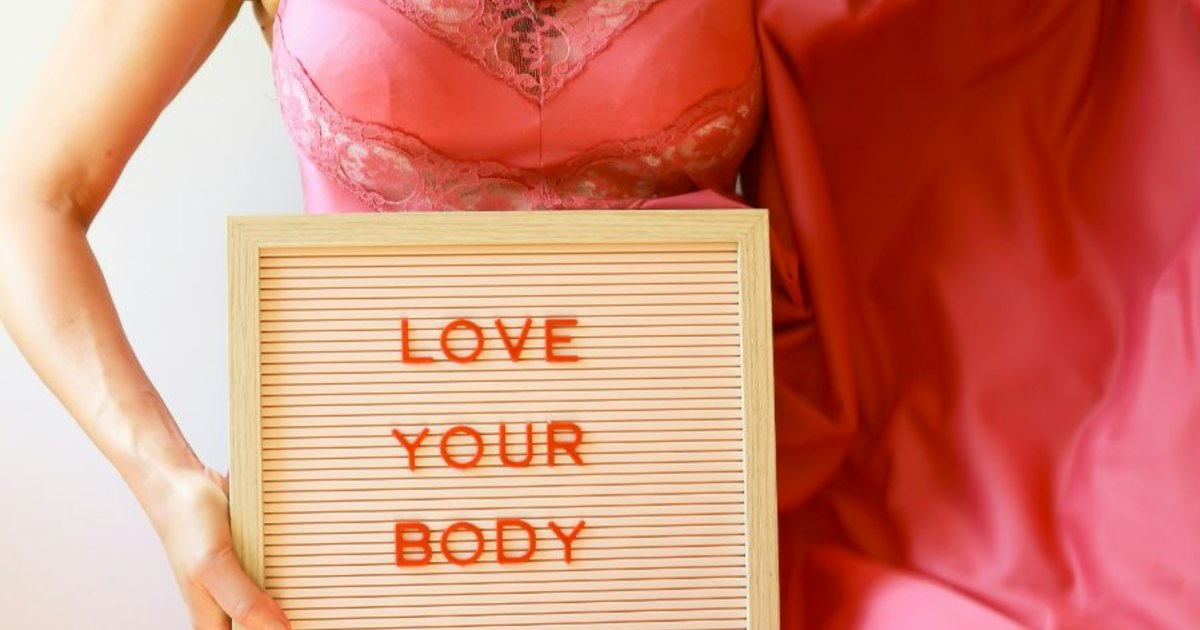 A person in a pink dress holding a letterboard reading LOVE YOUR BODY.