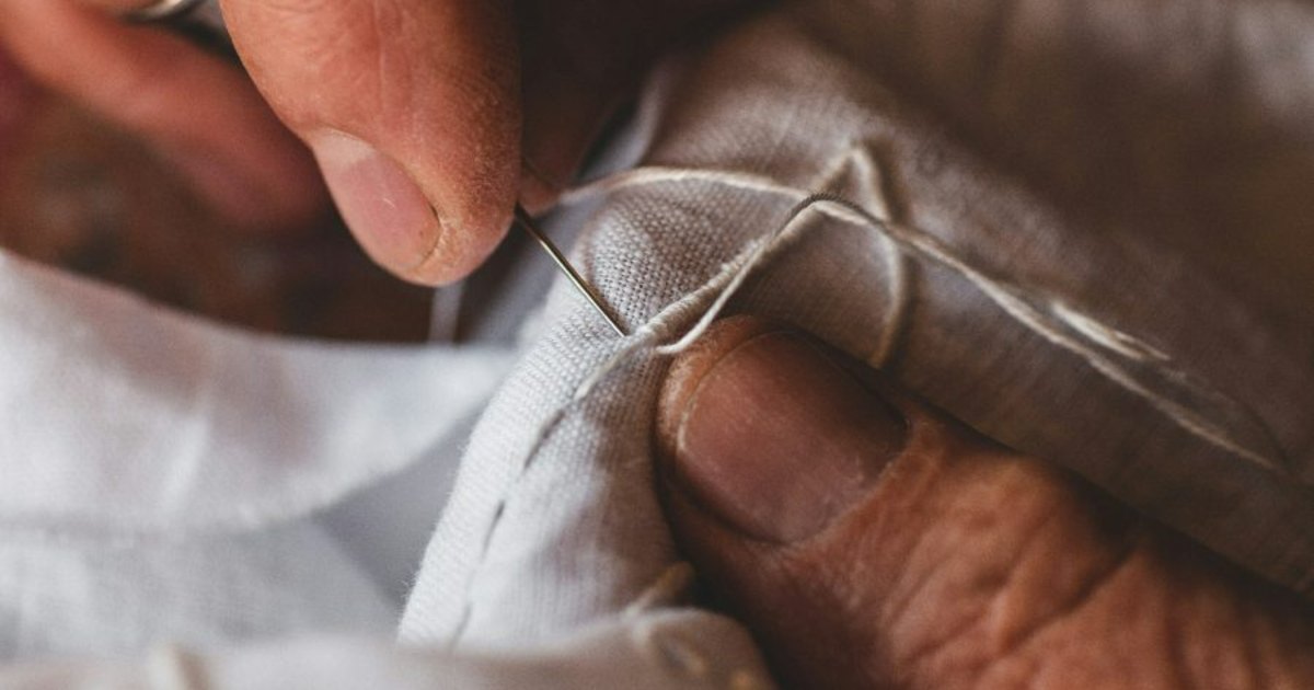 Close-up of hands sewing a neat repair into pale linen with a needle and thread.